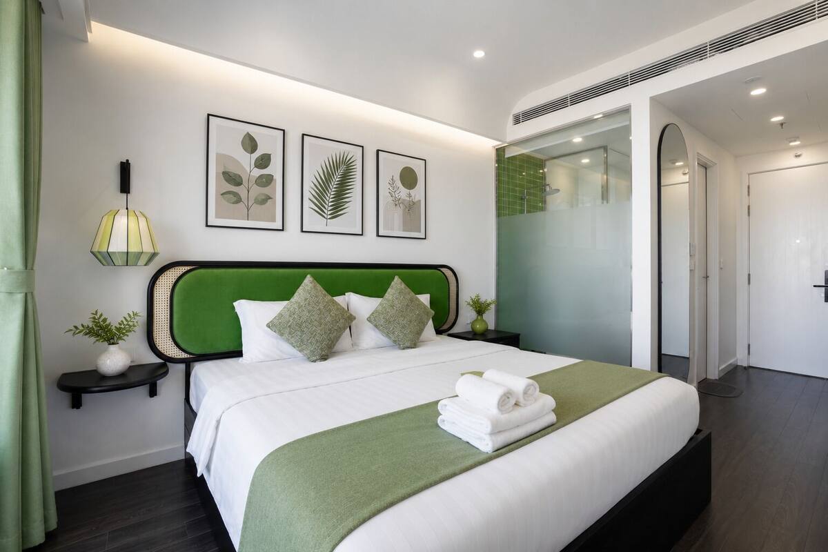 Un moderno cuarto de hotel con un cabecero tapizado en verde, una cama bien hecha con ropa blanca y detalles en verde, una mesita de noche con lámpara e impresiones botánicas enmarcadas en la pared.