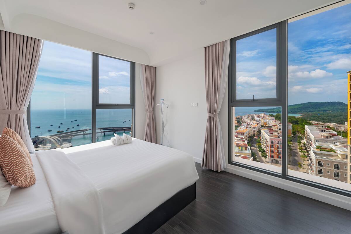 Habitación de hotel moderna con una cama grande, vistas al mar y a la ciudad, y un balcón con barandilla.