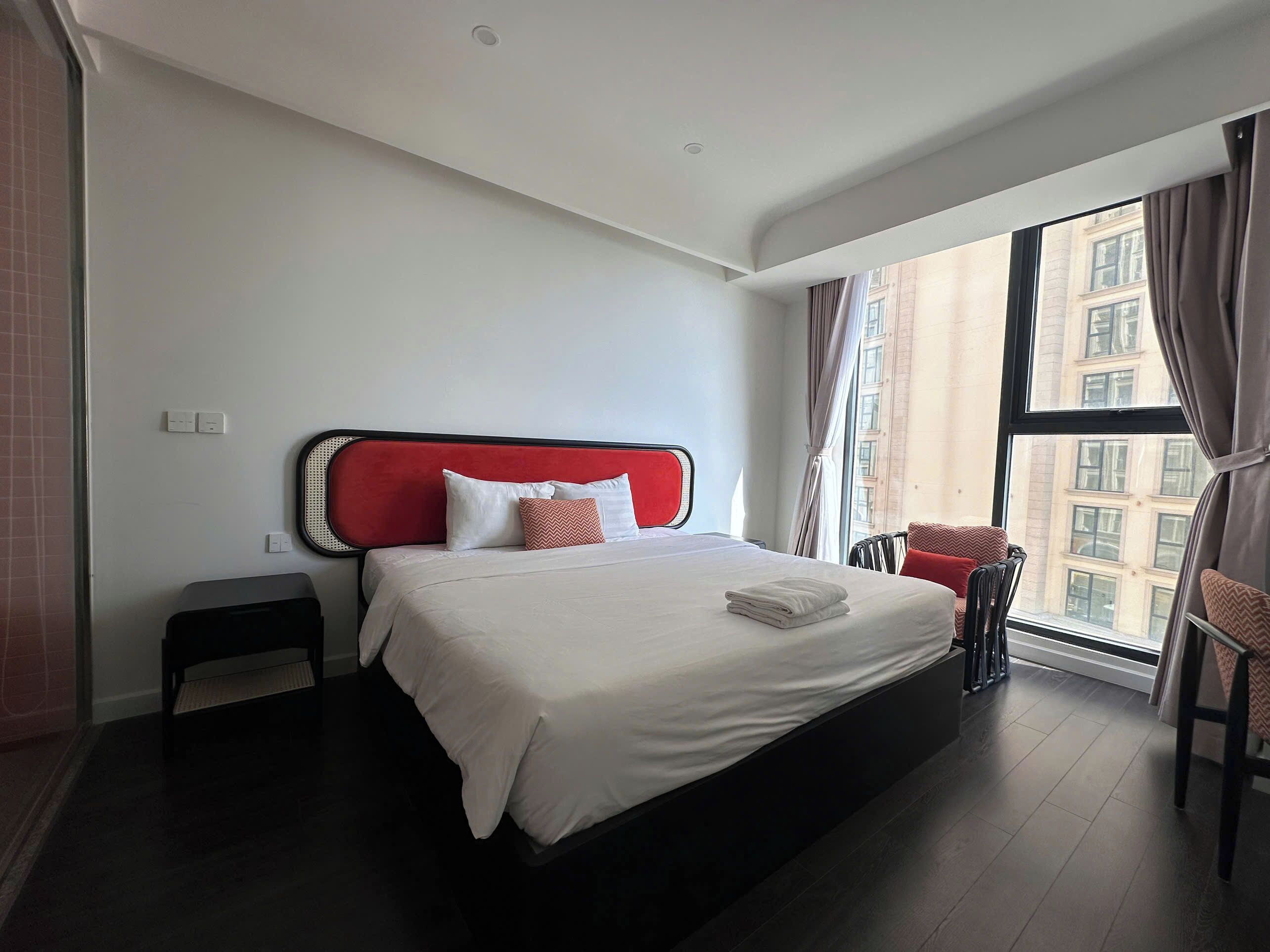 Habitació d'hotel moderna amb un llit gran, una petita taula auxiliar, una cadira i vista a la ciutat a través d'una finestra ampla.