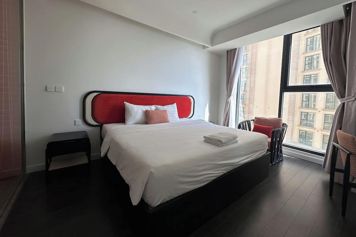 Una habitación de hotel moderna con una cama grande, una mesita auxiliar pequeña, una silla y vistas de la ciudad a través de un ventanal amplio.