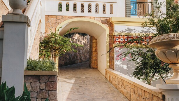 A pathway leading to a building with a blue facade, an arched entrance, and a decorative fountain. - Dido Carthage Apartment (An Thoi)