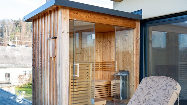 Een houten sauna met glazen deuren, een bank binnen en een stoel buiten.