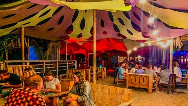 En restaurang vid stranden med träbord och träbänkar, uteservering och ett färgglatt parasoll som ger skugga.