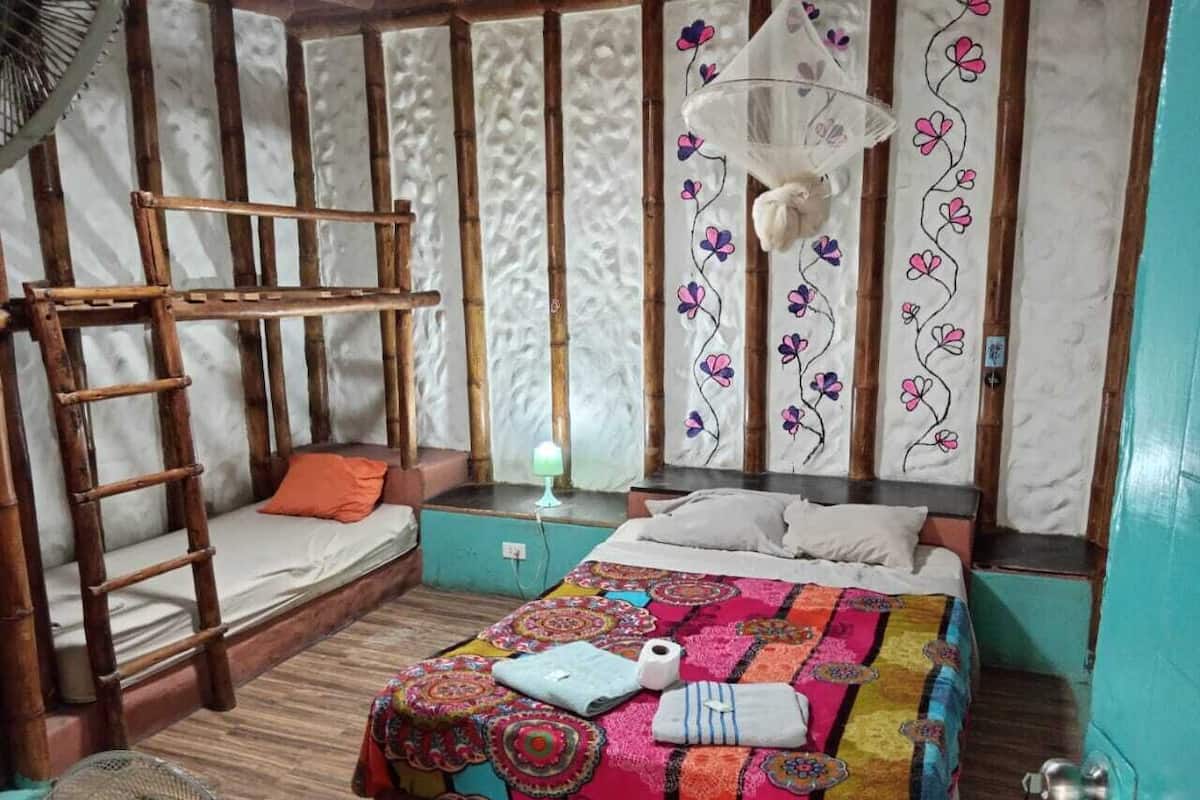 A room with a bunk bed, a bedside table with a lamp, a colorful quilt, and a fan.