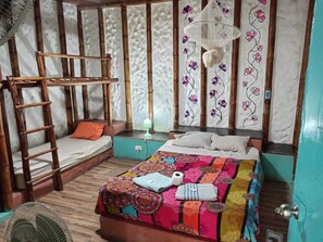 A room with a bunk bed, a bedside table with a lamp, a colorful quilt, and a fan.