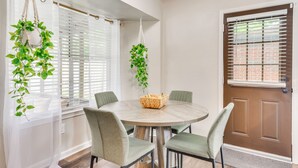 Un coin repas avec une table ronde, quatre chaises, un panier de fruits et deux plantes suspendues.