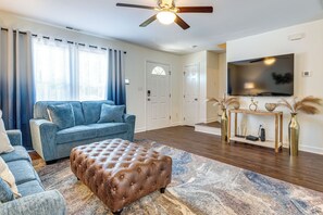 Ein Wohnzimmer mit einem blauen Sofa, einem braunen Ottomanen, einem Holzkonsolentisch mit Dekoration und einem angebrachten Fernseher.