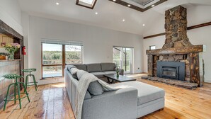 Uma sala de estar moderna com lareira de pedra, sofá seccional cinza, piso de madeira e grandes janelas com vista para o exterior.