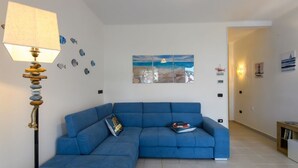 Una sala de estar con un sofá seccional azul, una lámpara con pantalla texturizada, una alfombra y obras de arte que representan una escena de playa.