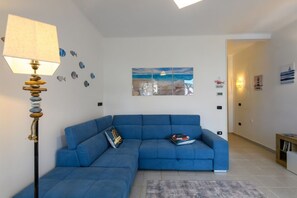 Ein Wohnzimmer mit einem blauen Ecksofa, einer Lampe mit strukturierter Schirm, einem Teppich und Wandbildern, die eine Strandansicht zeigen.