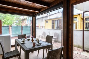 Eine überdachte Terrasse mit Esstisch, Korbstühlen und Blick auf benachbarte Gebäude.