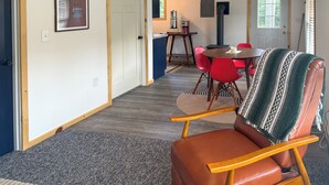 Huoneessa on ruskea nahkainen nojatuoli, punaisilla tuoleilla varustettu ruokapöytä ja kuviollinen matto.
