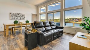 Uma sala de estar espaçosa com sofá de couro, mesa de jantar com cadeiras e janelas amplas com vista para um corpo de água e edifícios.