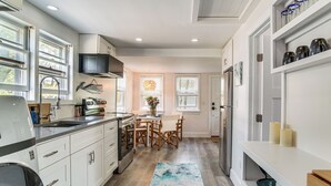 A modern kitchen with white cabinetry, a central island, and a dining area with wooden chairs. - Periwinkle: Gateway to Cape Cod Coastal Cottage (Wareham)