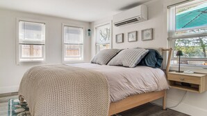 A bedroom with a bed, bedside table, lamp, air conditioning unit, and window blinds. - Periwinkle: Gateway to Cape Cod Coastal Cottage (Wareham)