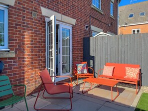 Ein Ziegelhaus mit Schiebetür, einem Holzzaun und Gartenmöbeln, darunter ein grüner Sessel, ein orangefarbenes Sofa und ein passender Tisch.