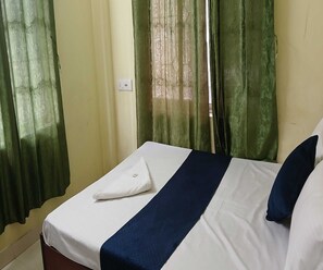 Een eenpersoonsbed met een wit- en blauwe sprei, groene gordijnen en een zichtbare stopcontact op de muur.