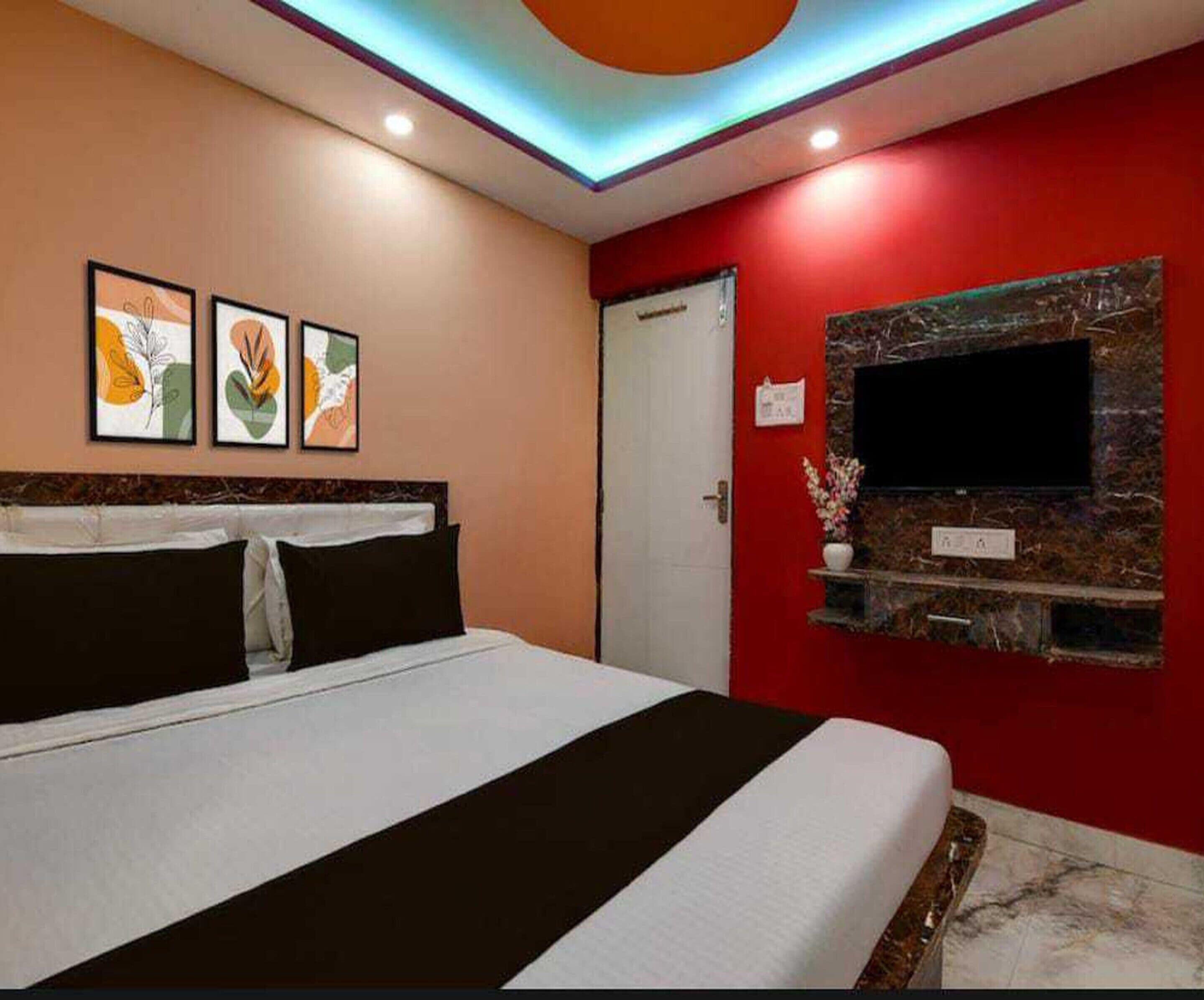 A hotel room with a bed, a TV mounted on a red wall, and three framed artworks above the bed.