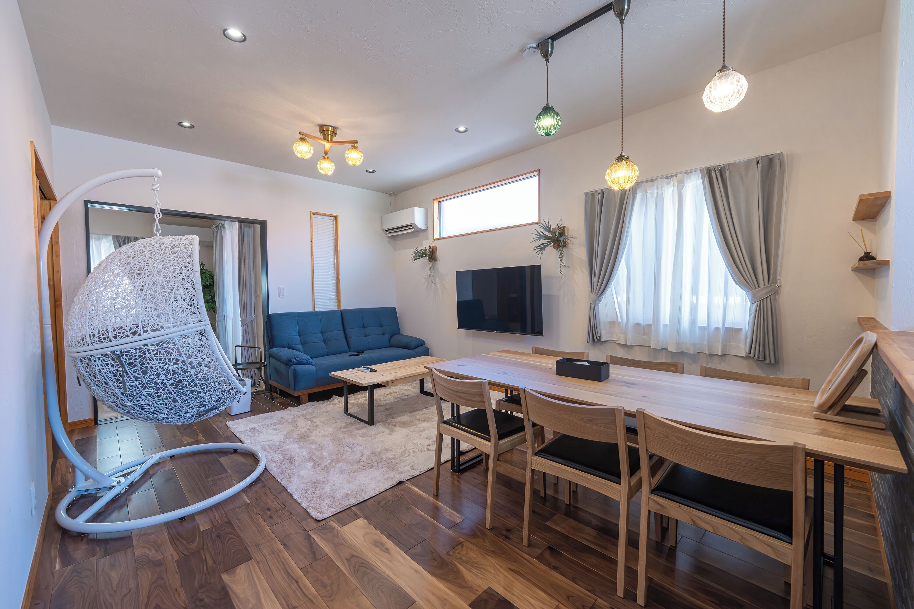 A modern living room with a blue sofa, a dining table with chairs, a hanging chair, a flat-screen TV, and a large window with curtains.
