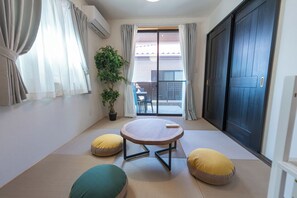 A room with tatami flooring, a low table, cushions, a potted plant, and a sliding glass door leading to an outdoor area. - Miyako Island Breeze (Miyakojima City)