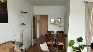 Ein Wohnzimmer mit Sofa, einem Sessel, einem Esstisch mit Stühlen, einer Lampe, einer Zimmerpflanze und einem gerahmten Bild an der Wand.