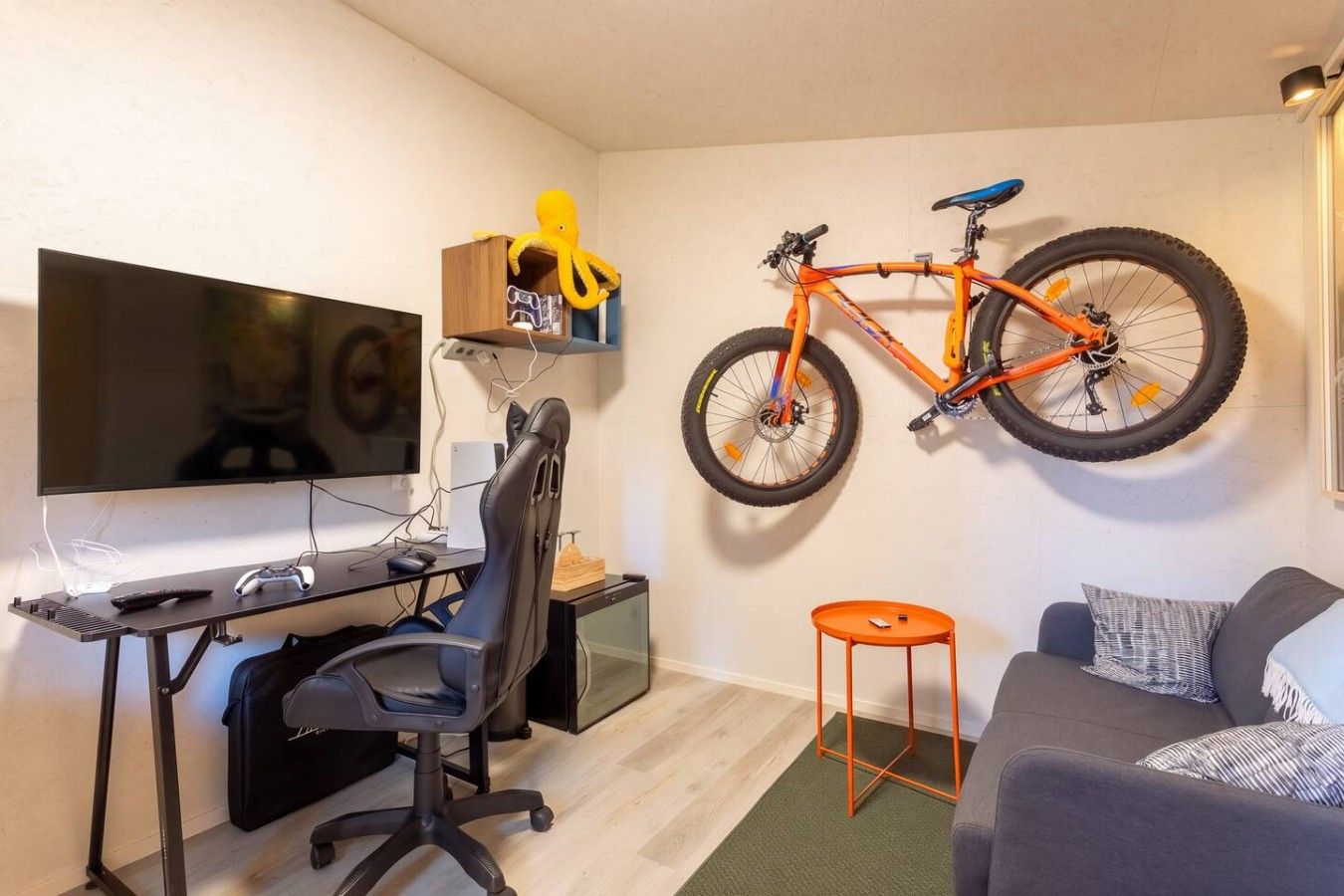 Una habitación con televisor, silla gamer, escritorio, bicicleta naranja montada en la pared y un sofá.