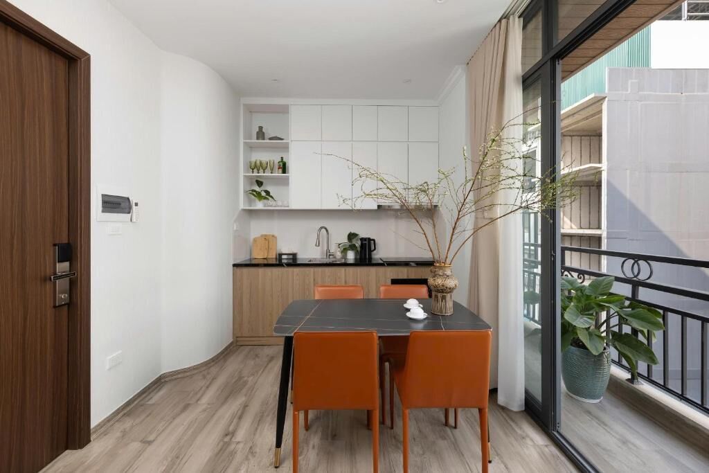 A dining area with a table and chairs, a kitchen in the background, and a balcony with a railing and a potted plant.