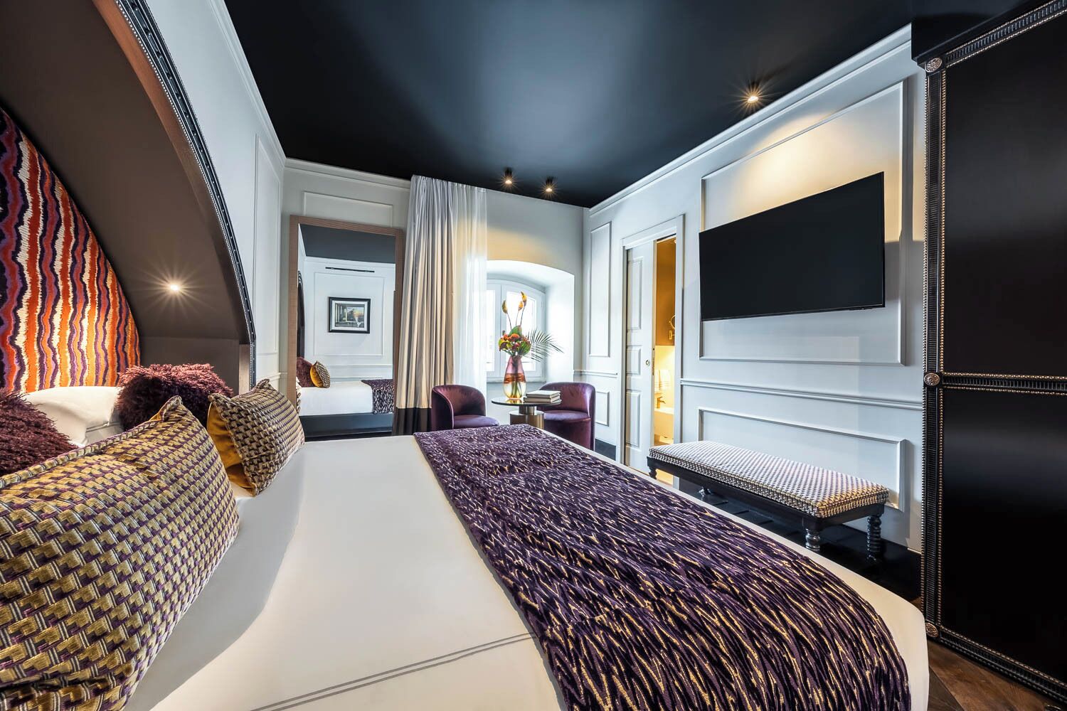 Quarto de hotel moderno com uma cama grande, área de estar, televisão e vista para o corredor.
