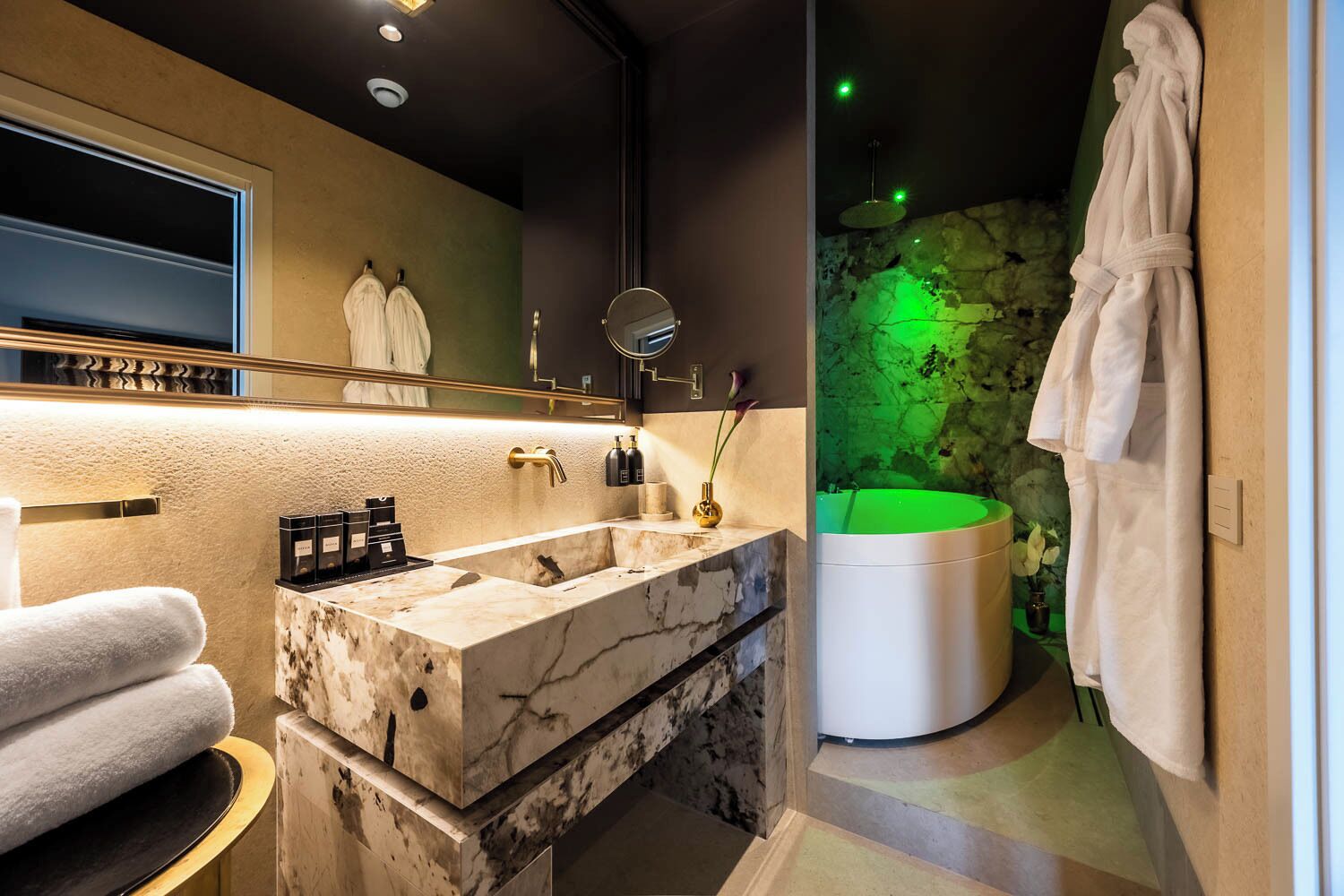 Un baño moderno con lavamanos de mármol, un espejo grande, una pared iluminada en verde y una bañera.