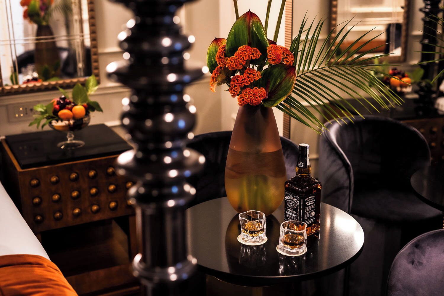 A round table with a vase of flowers, a bottle of whiskey, and two glasses.