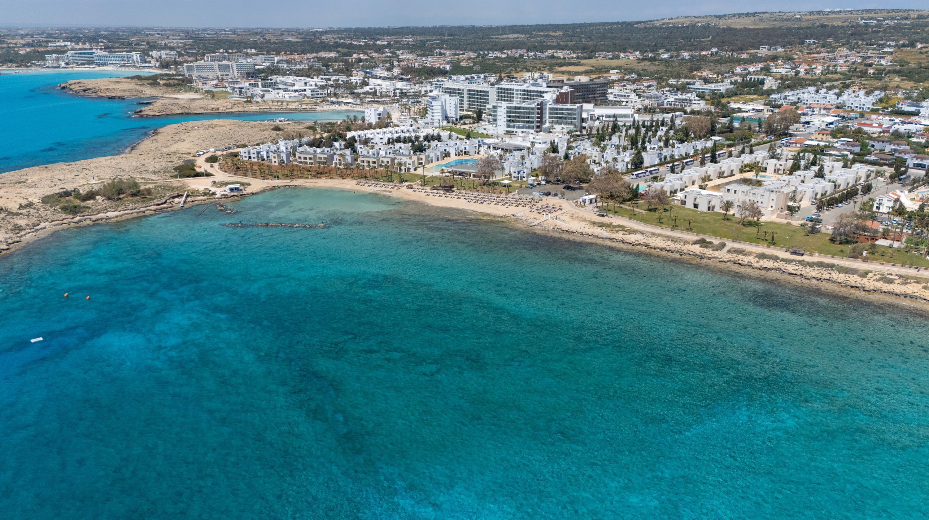 Vista aérea de una zona turística costera con edificios, una playa y aguas azules cristalinas.