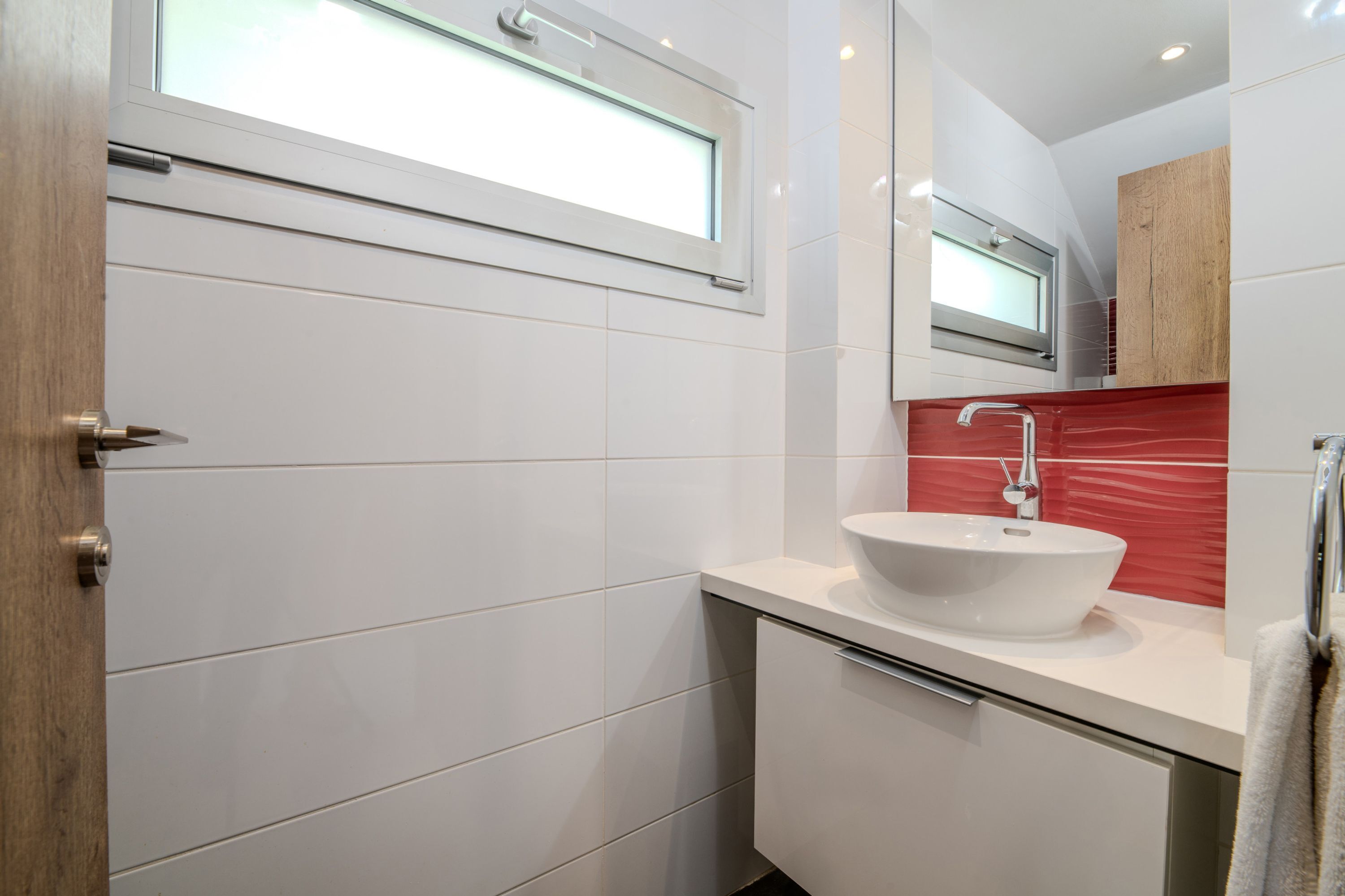 A modern bathroom with a white tiled wall, a large window, a white sink, and a red-tiled backsplash.