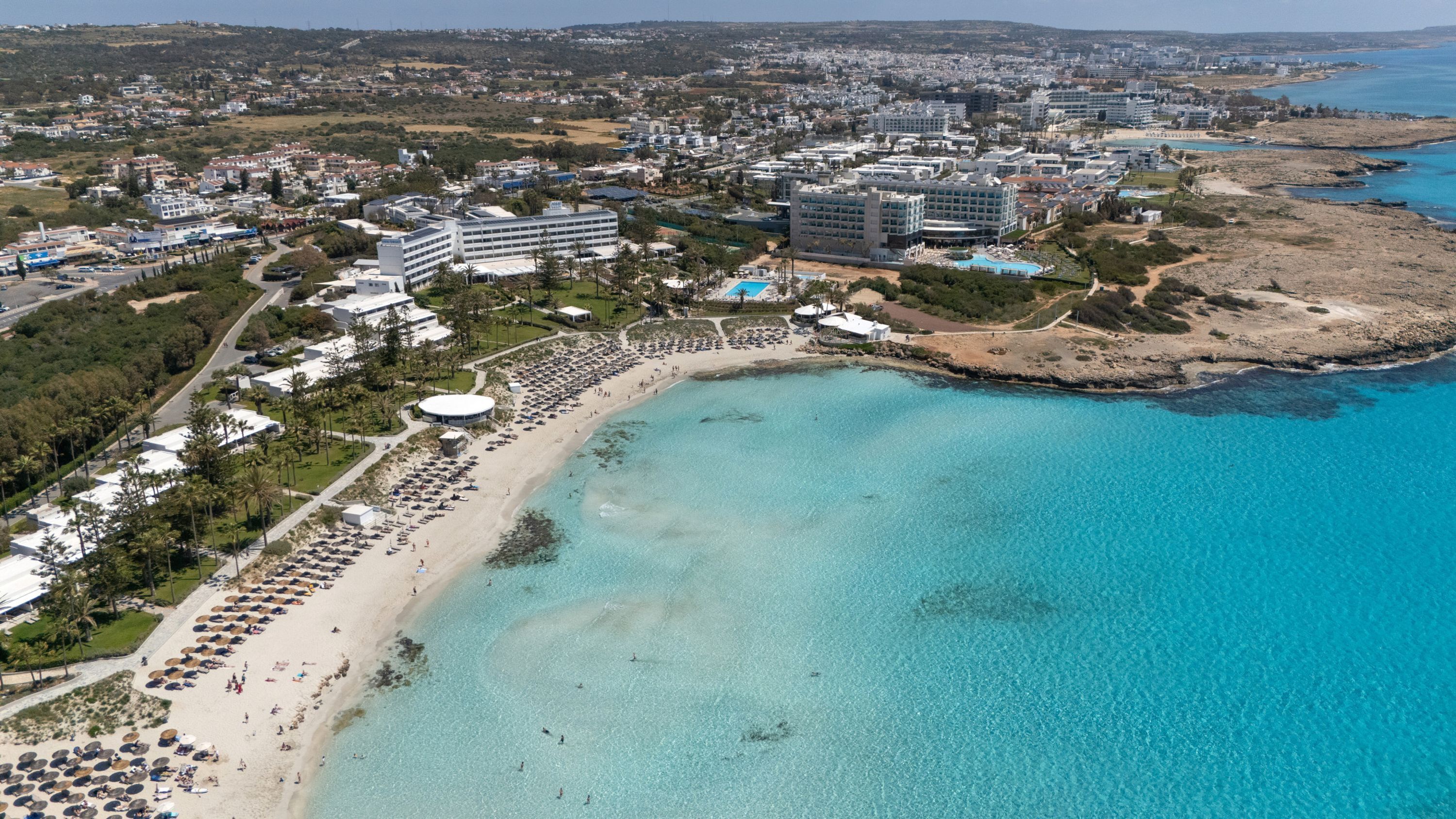 Vista aérea de un complejo turístico frente al mar, con una playa, varios edificios de hotel y un mar azul cristalino.