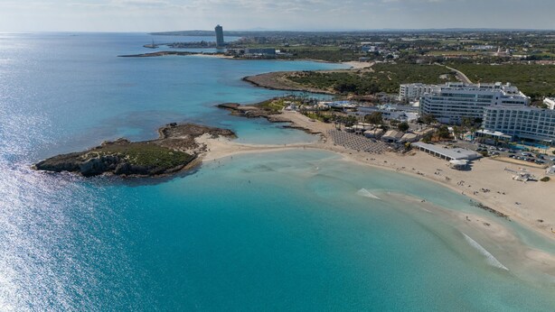 Ett flygbildsbild av ett kustområde med en strand, en småbåtshamn och ett hotellkomplex.