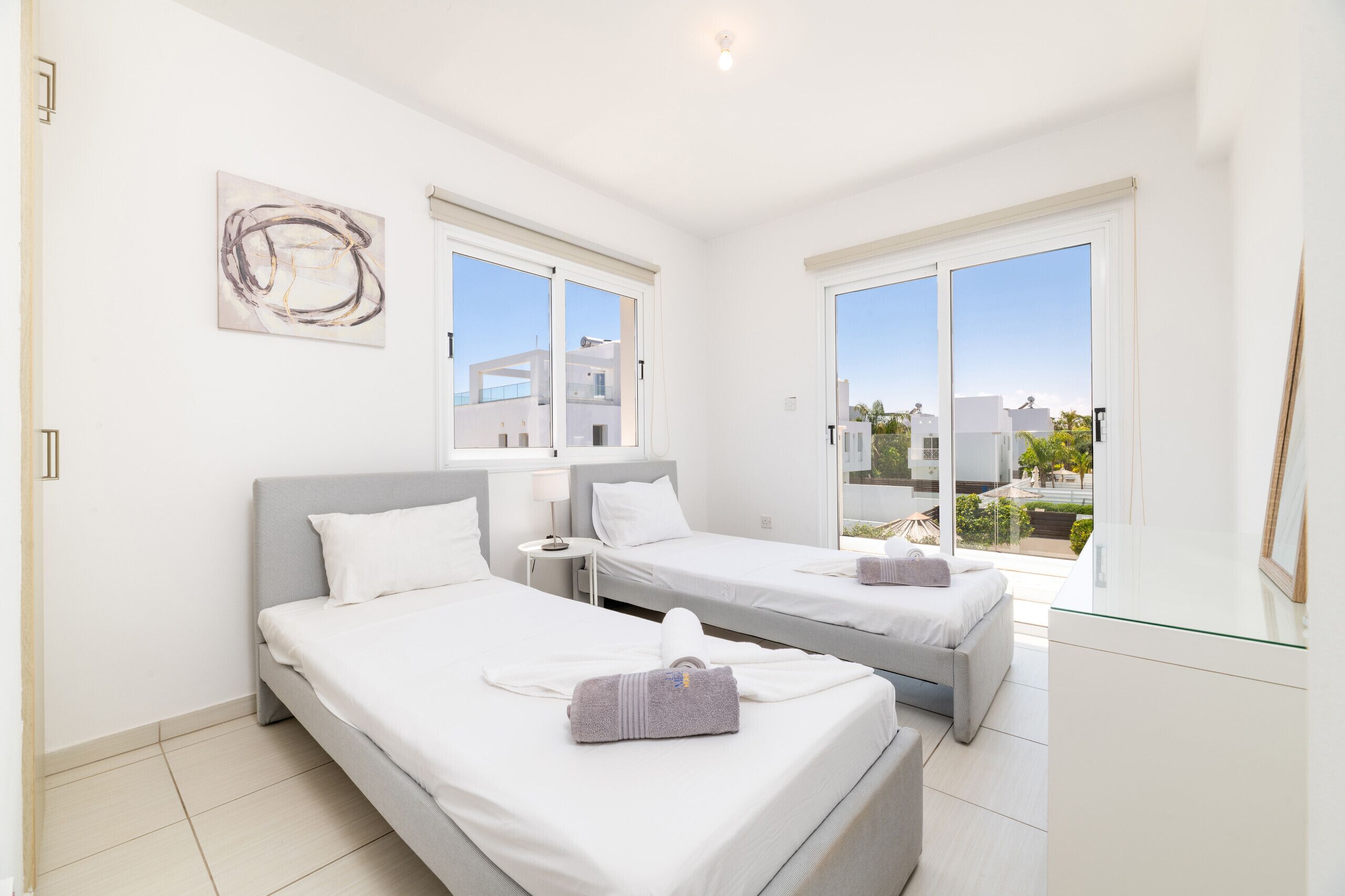 A modern hotel room with two beds, a glass door leading to a balcony, and a view of the cityscape.