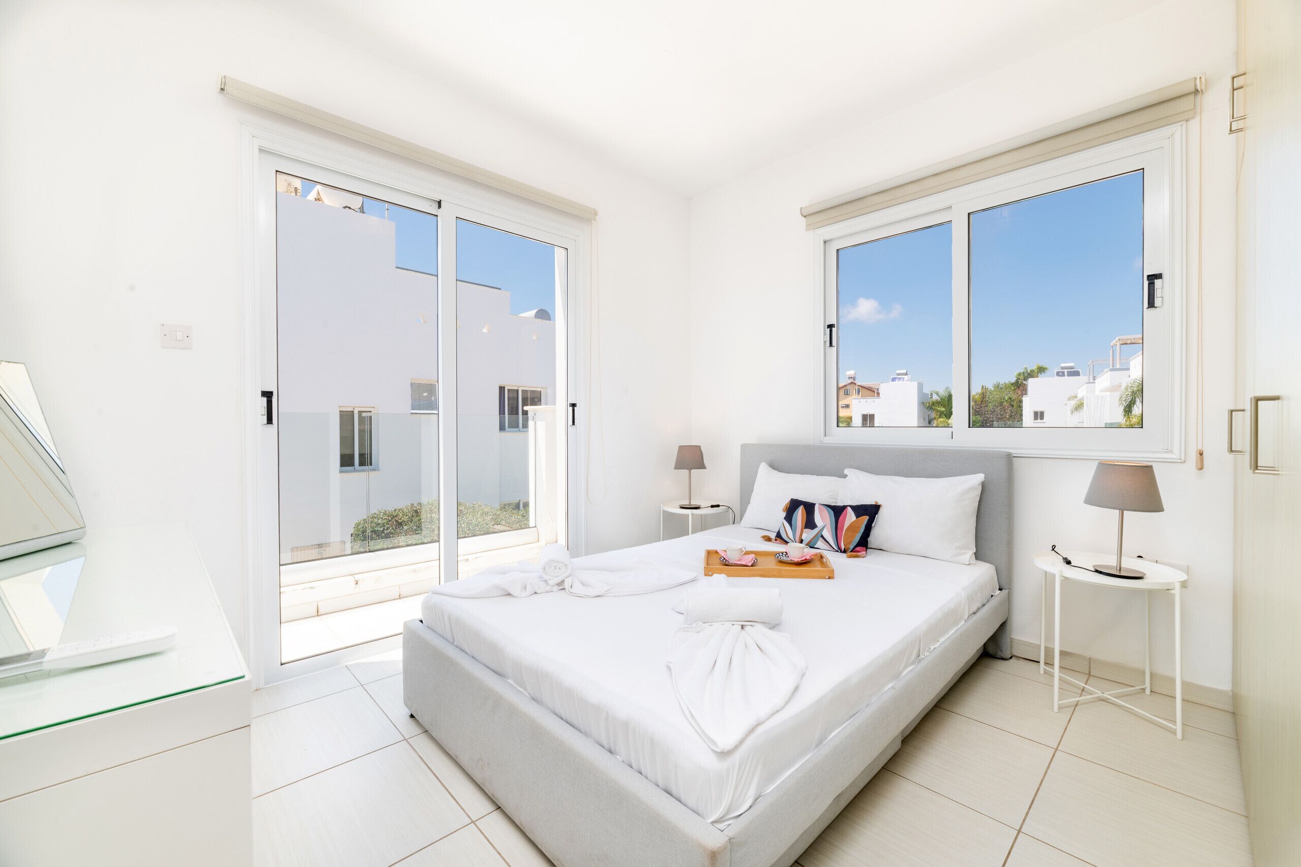 A modern bedroom with a bed, two bedside tables, a glass desk, and large windows.