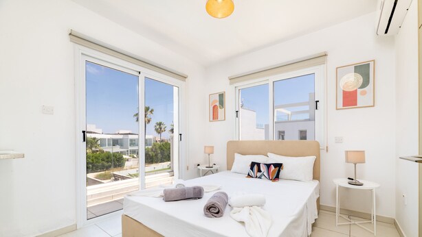 A modern bedroom with a large bed, two bedside tables, a view through a sliding door, and artwork on the walls. - Nissini Villa 15 in Ayia Napa (Ayia Napa)