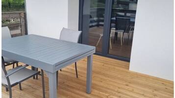 Un patio extérieur moderne avec une table et des chaises grises, un plancher en bois et une porte vitrée donnant sur une terrasse.