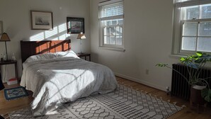 Ein Schlafzimmer mit Bett, Nachttisch, Lampe, einem gerahmten Bild, einem Fenster und einer Heizung.