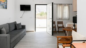A modern room with a grey sofa, a TV mounted on the wall, a dining table with chairs, and a sliding glass door leading to an outdoor area. - Black Sand Santorini Studio - 20m to Perissa Beach (Perissa)
