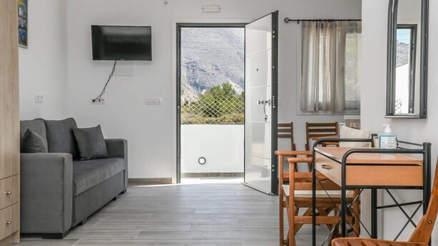 A modern room with a grey sofa, a TV mounted on the wall, a dining table with chairs, and a sliding glass door leading to an outdoor area with a mountain view. - Black Sand Santorini Studio - 20m to Perissa Beach (Perissa)