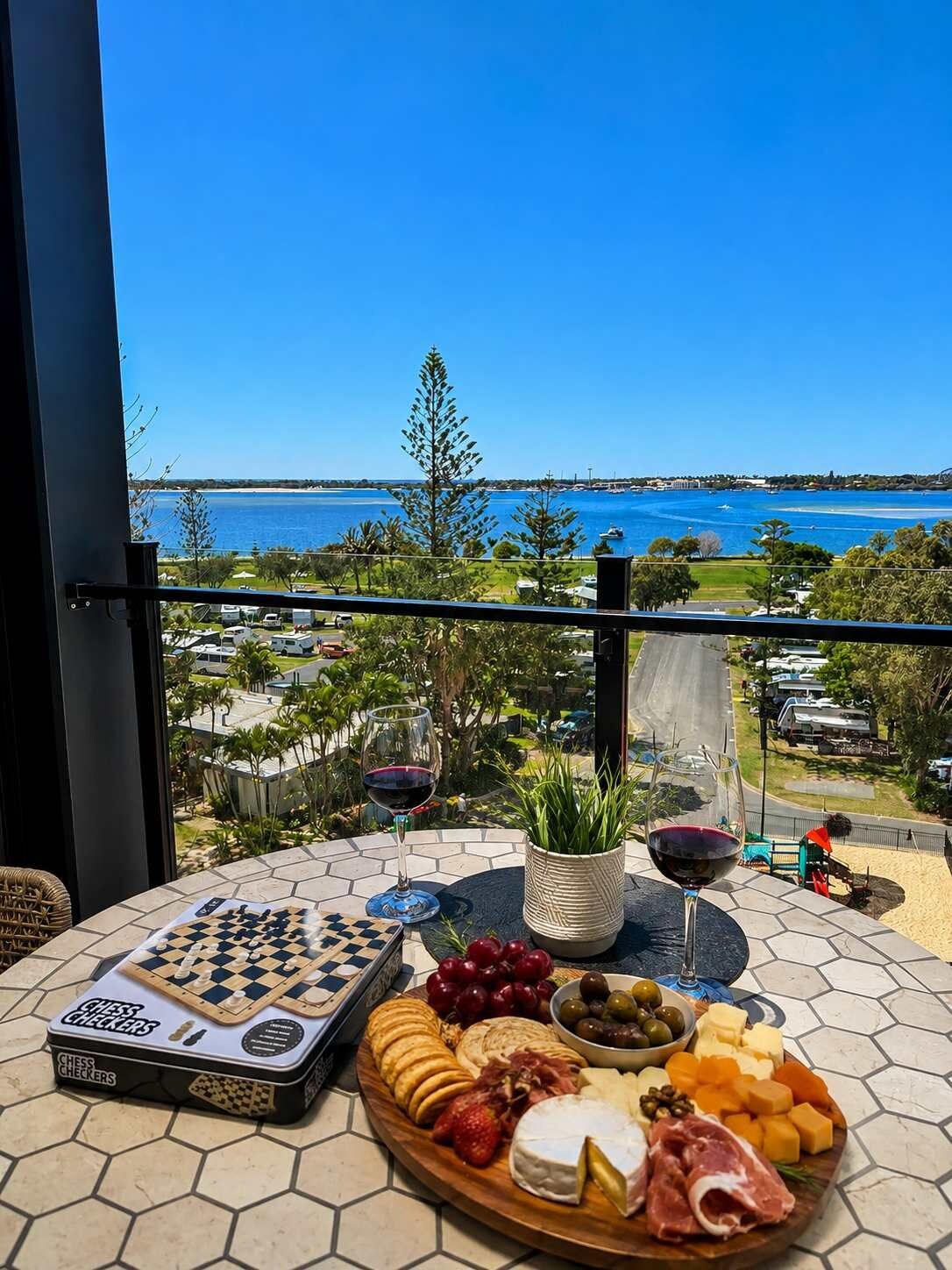 Un jeu d’échecs, des verres à vin et une planche de charcuterie garnie de différents fromages et de différentes sortes de viande, le tout disposé sur un balcon offrant une vue sur la côte.
