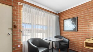 A room with brick walls, a white door, a window with sheer curtains, two black leather chairs, a small table, and a framed picture on the wall. - Edithburgh Seaside Motel (Edithburgh)