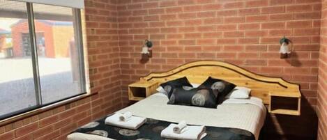 Un dormitorio con pared de ladrillo, cabecera de madera, cama con almohadas y colcha, y una ventana.
