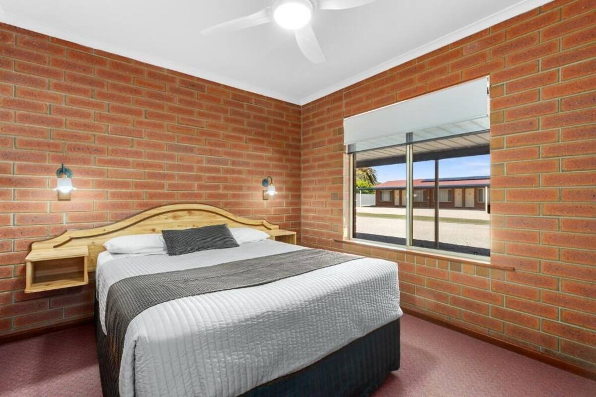 Un dormitorio con pared de ladrillo, cama con cabecera de madera, ventilador de techo y ventana con vistas al exterior.