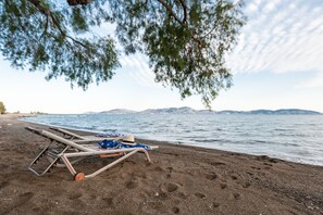 Ein Strand mit Liegestuhl und Hut im Sand, ein ruhiges Meer und Berge im Hintergrund.