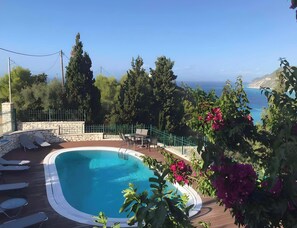 Ein Pool mit Liegestühlen, umgeben von viel Grün und Blick auf das Meer.