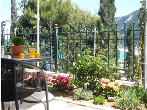 Ein Balkon mit Tisch, Topfpflanze und Getränken mit Blick auf eine Küstenlandschaft mit Bäumen und einem Berg.