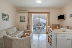 A living room with a sofa, a coffee table, a dining table with chairs, a TV mounted on the wall, and a balcony with a view. - Kyma Arch House: Authentic Stay with Panoramic Views in Porto Heli (Porto Cheli)