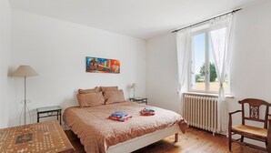 A bedroom with a bed, a chair, a table, a radiator, a window with curtains, and a painting on the wall. - Holiday Home 'Le Café De L'Encloître' with Private Terrace, Private Garden and Wi-Fi (Beaumont-Louestault)